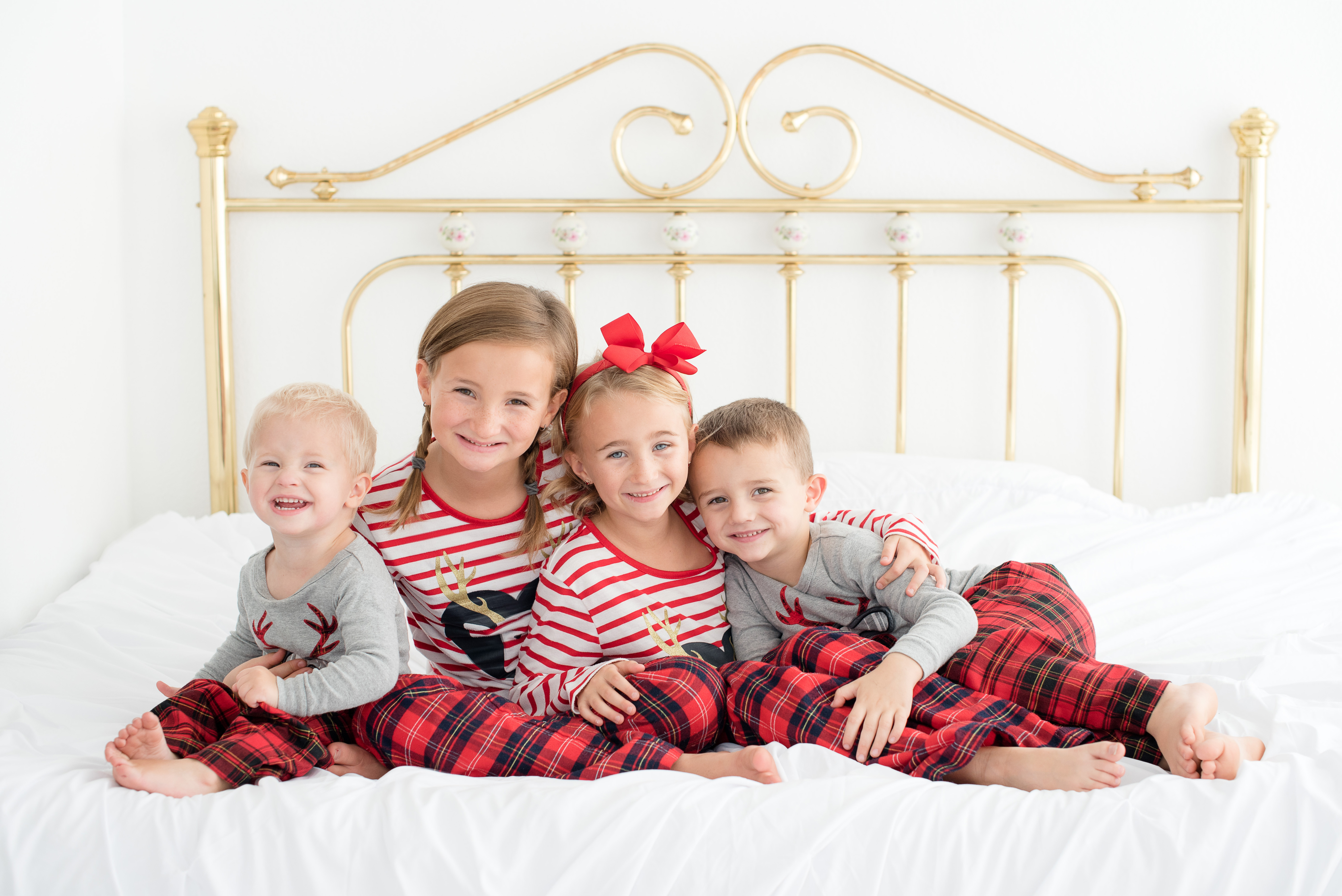 Color photograph of four children in Christmas pajamas sitting on a white bed with a brass headboard
